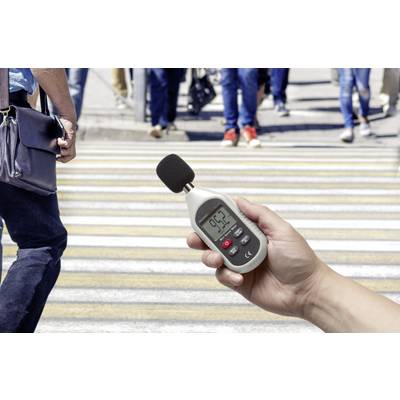 A hand is holding a sound level meter displaying 95.3 decibels. In the background, people are crossing a pedestrian crossing.