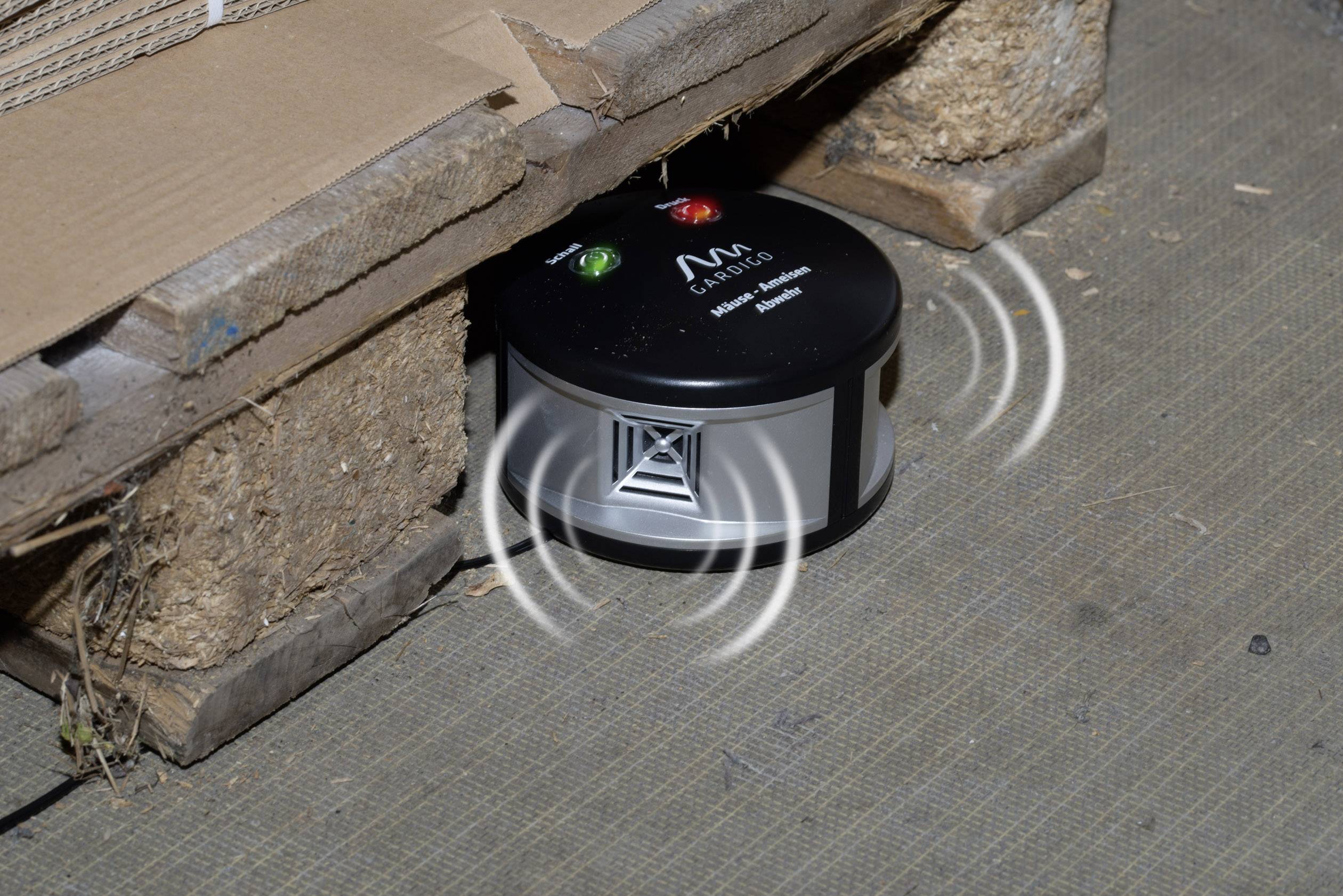 'Small black device with green and red indicator lights on the floor beneath a pallet, emitting sound waves.'
