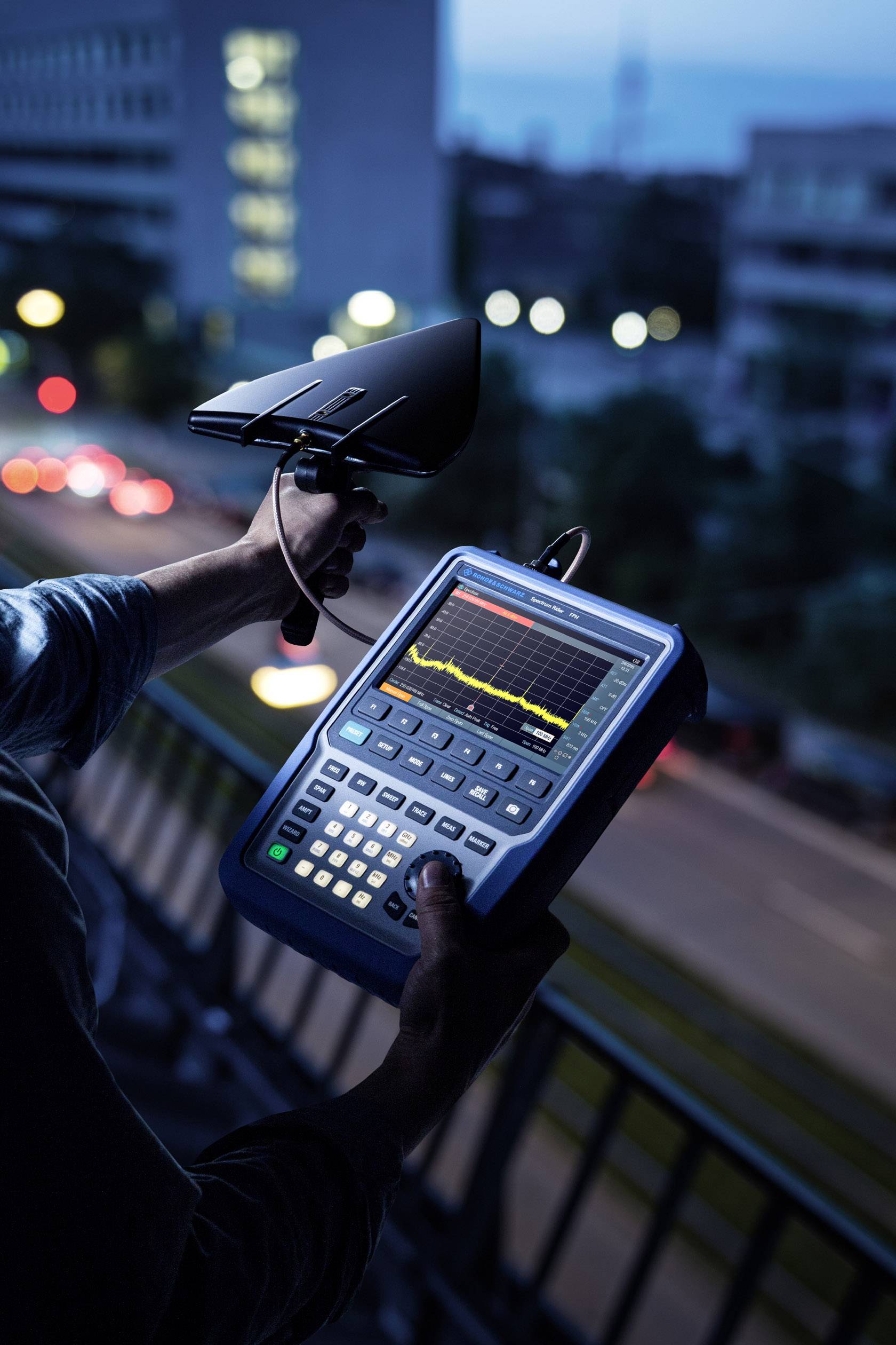 A person is holding a measuring device with a graphical display in a city at dusk, likely checking signals.