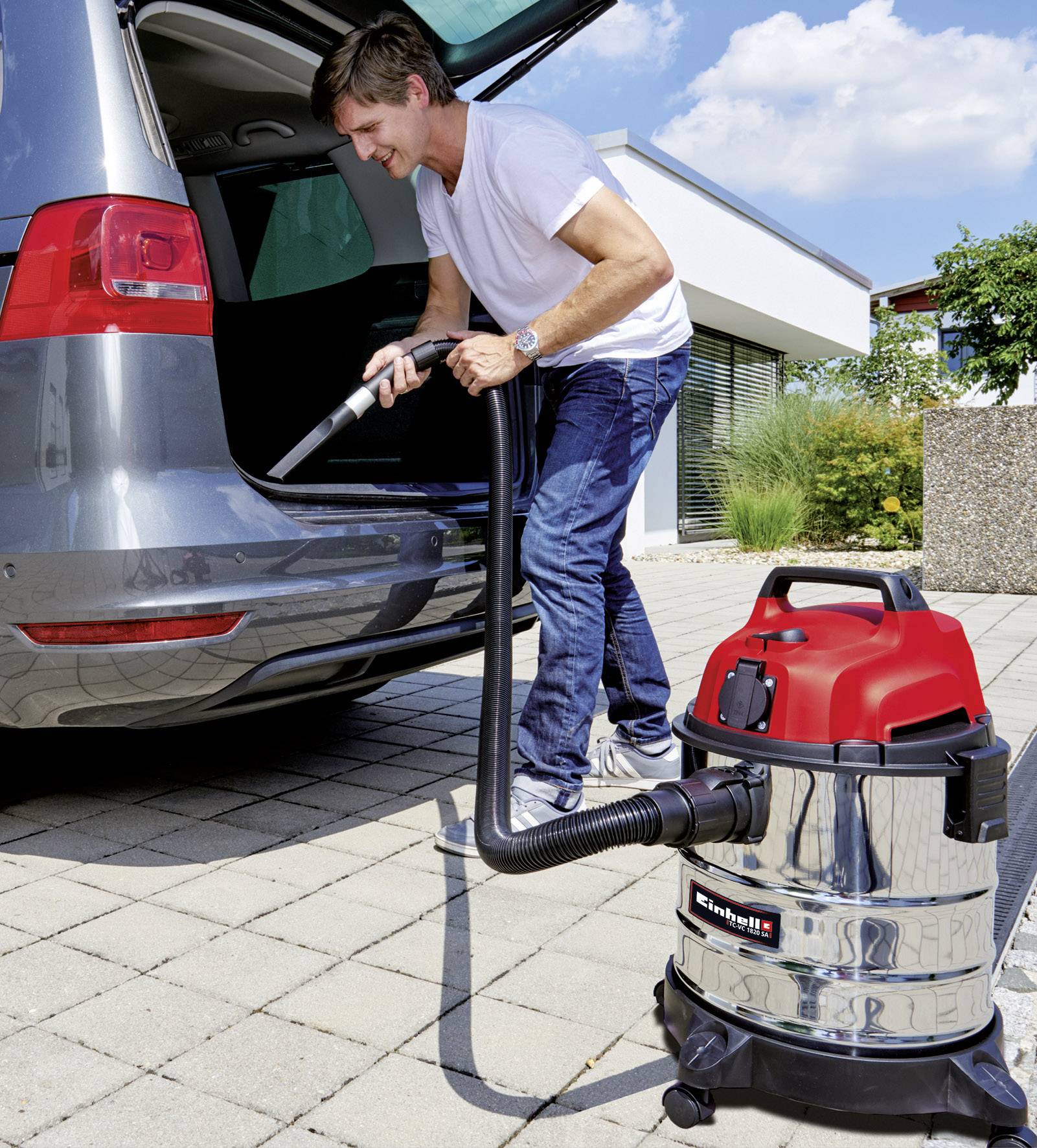 A man is cleaning the boot of a car with a wet and dry vacuum cleaner on a paved driveway in front of a modern house.