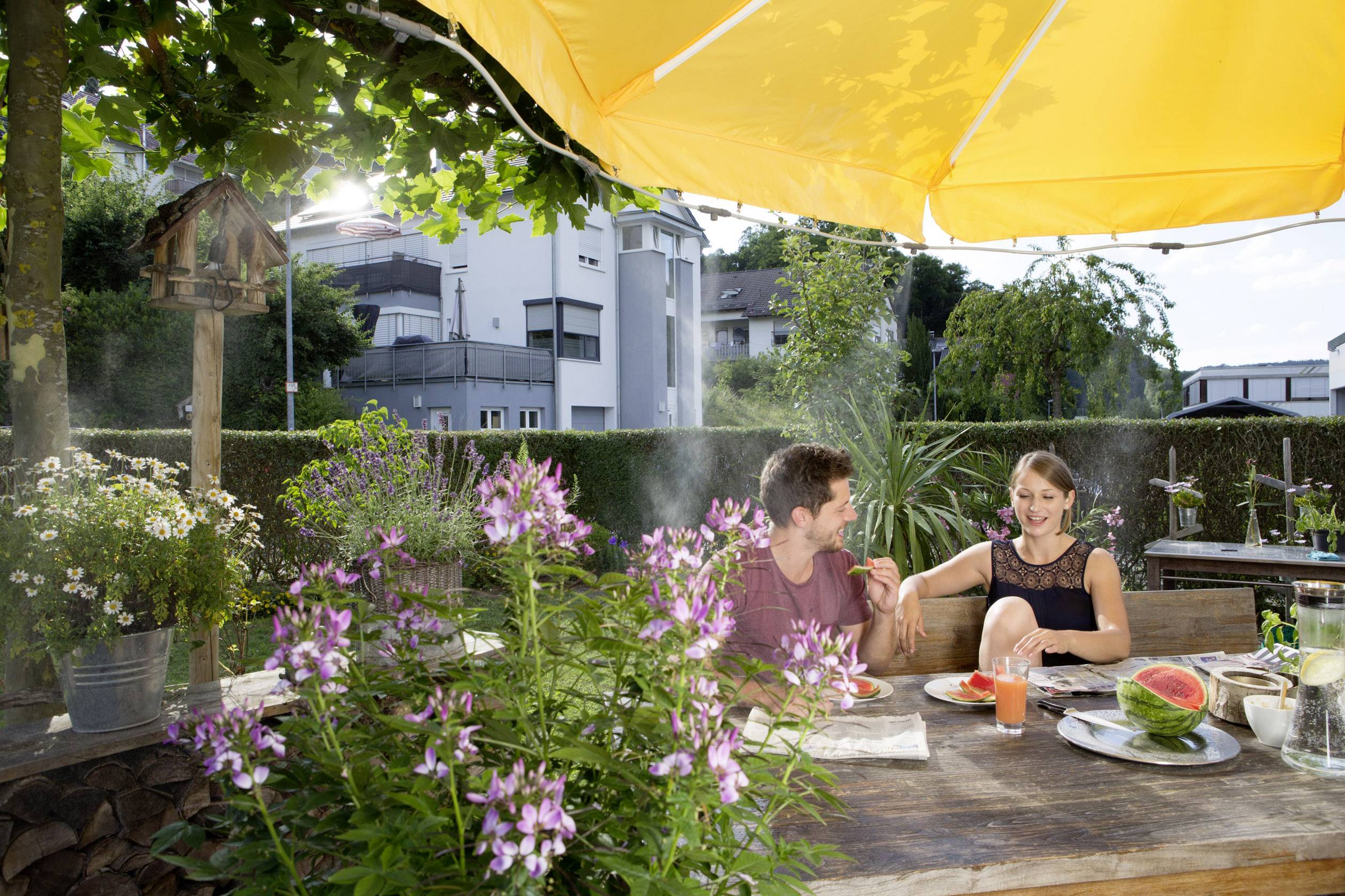 A couple sit under a yellow parasol in the garden, eating together at a wooden table. Flowers in the foreground, residential houses in the background.