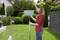 A woman is standing in the garden holding a blue broom. Various green plants and a fence can be seen in the background.