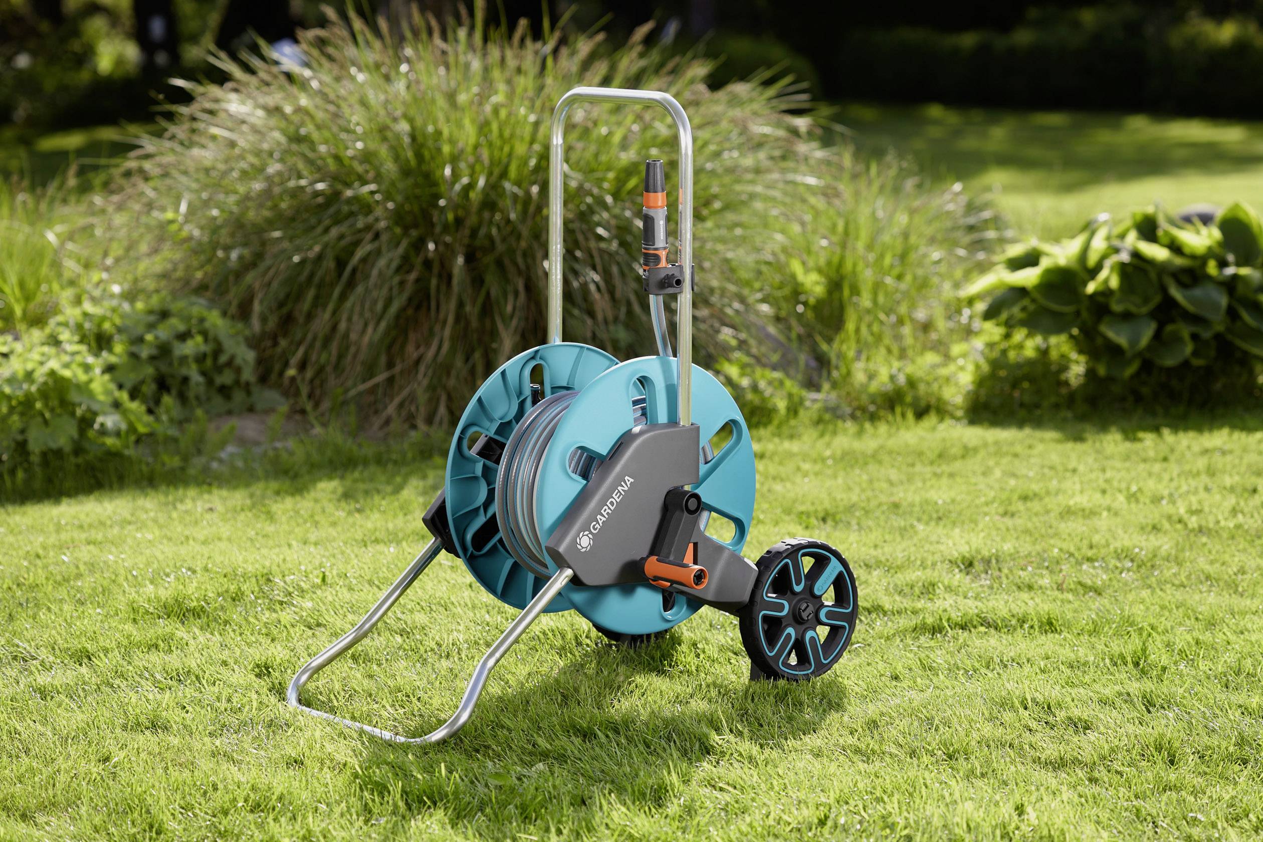 A garden hose reel sits on the lawn, featuring a turquoise hose reel and two black wheels, surrounded by plants.