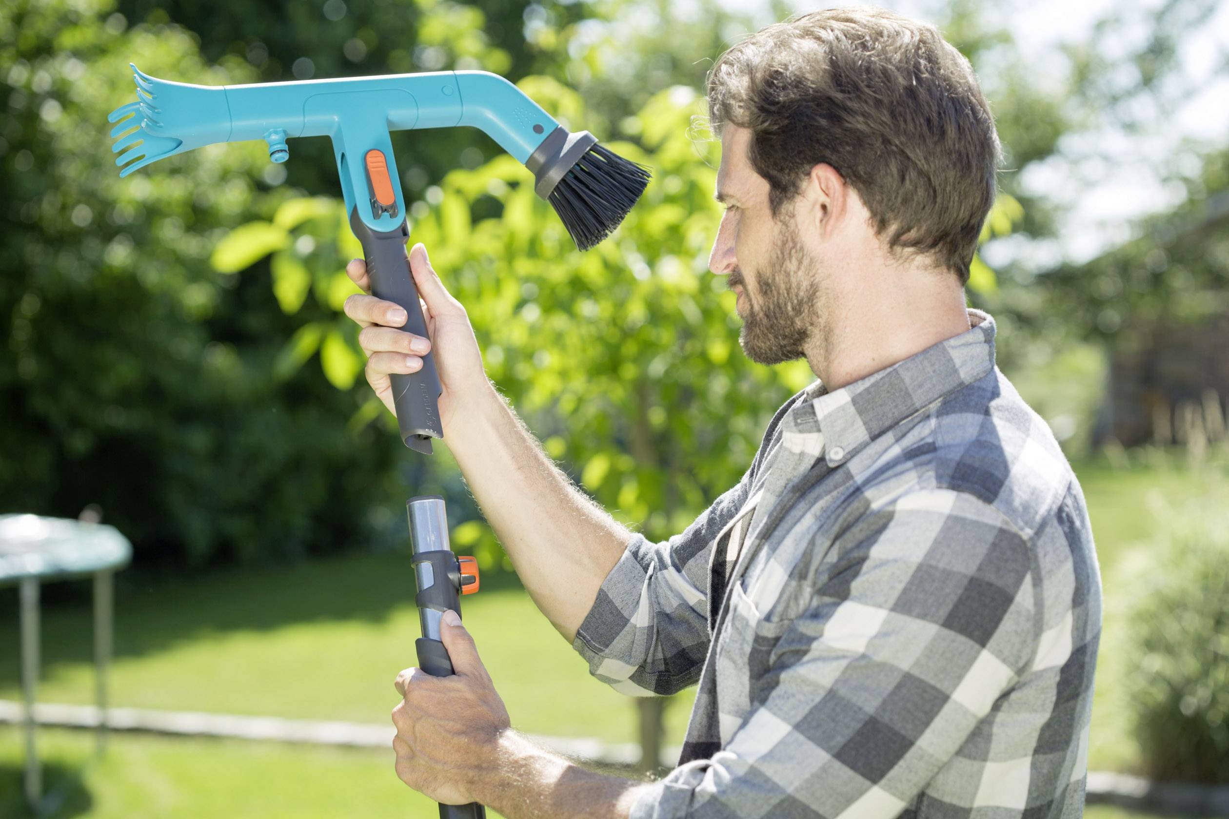 A man in a checked shirt is holding a gardening tool with a brush in his hand, with a green garden in the background.