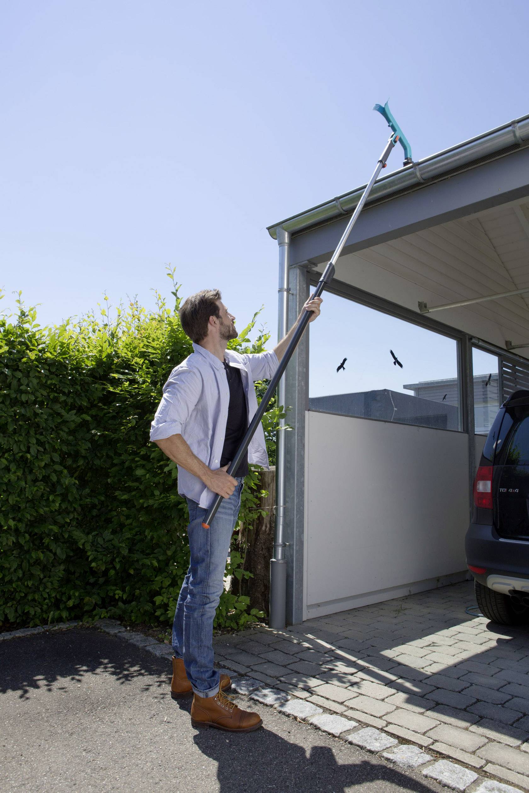 A man is cleaning an awning with a long brush next to a car. In the background, a green bush is visible under a clear sky.