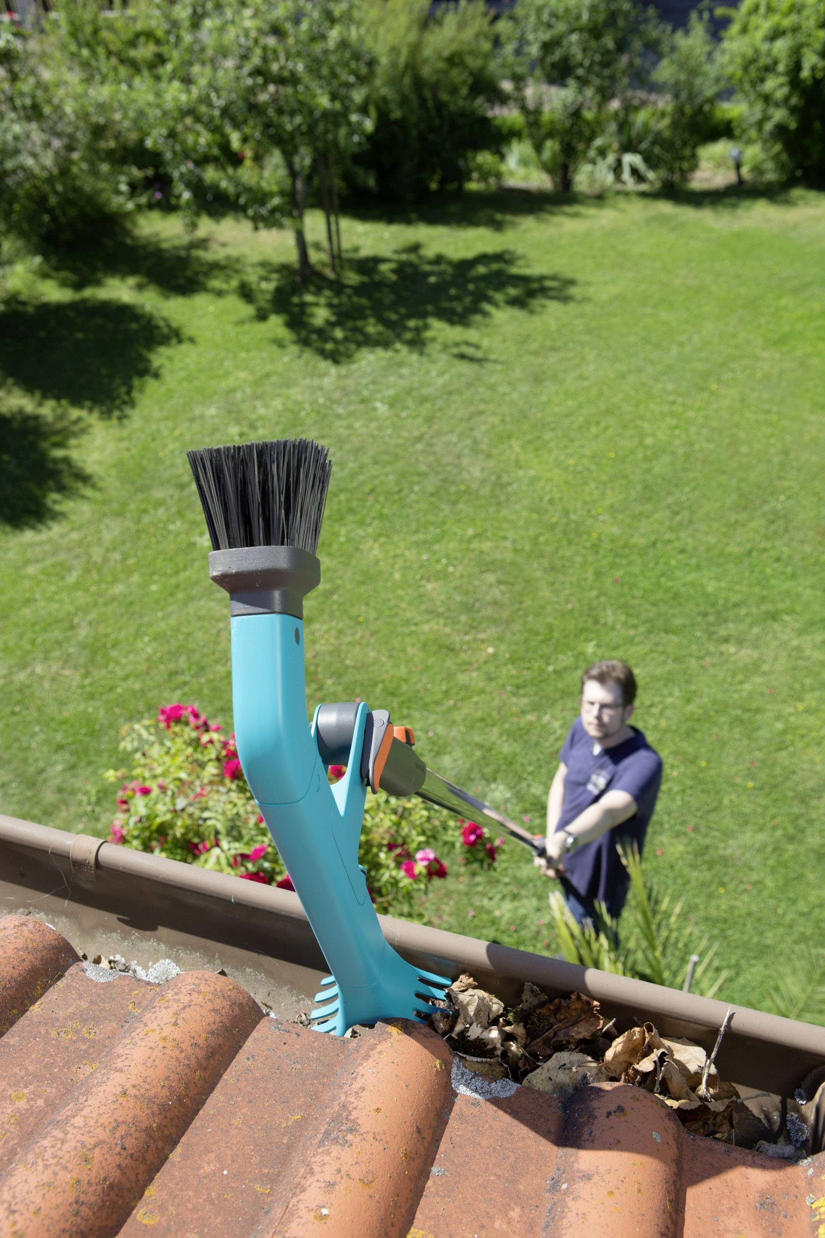 A person is cleaning a gutter with a blue brushing device. They are standing in the garden, with the brush positioned at the roof's edge.