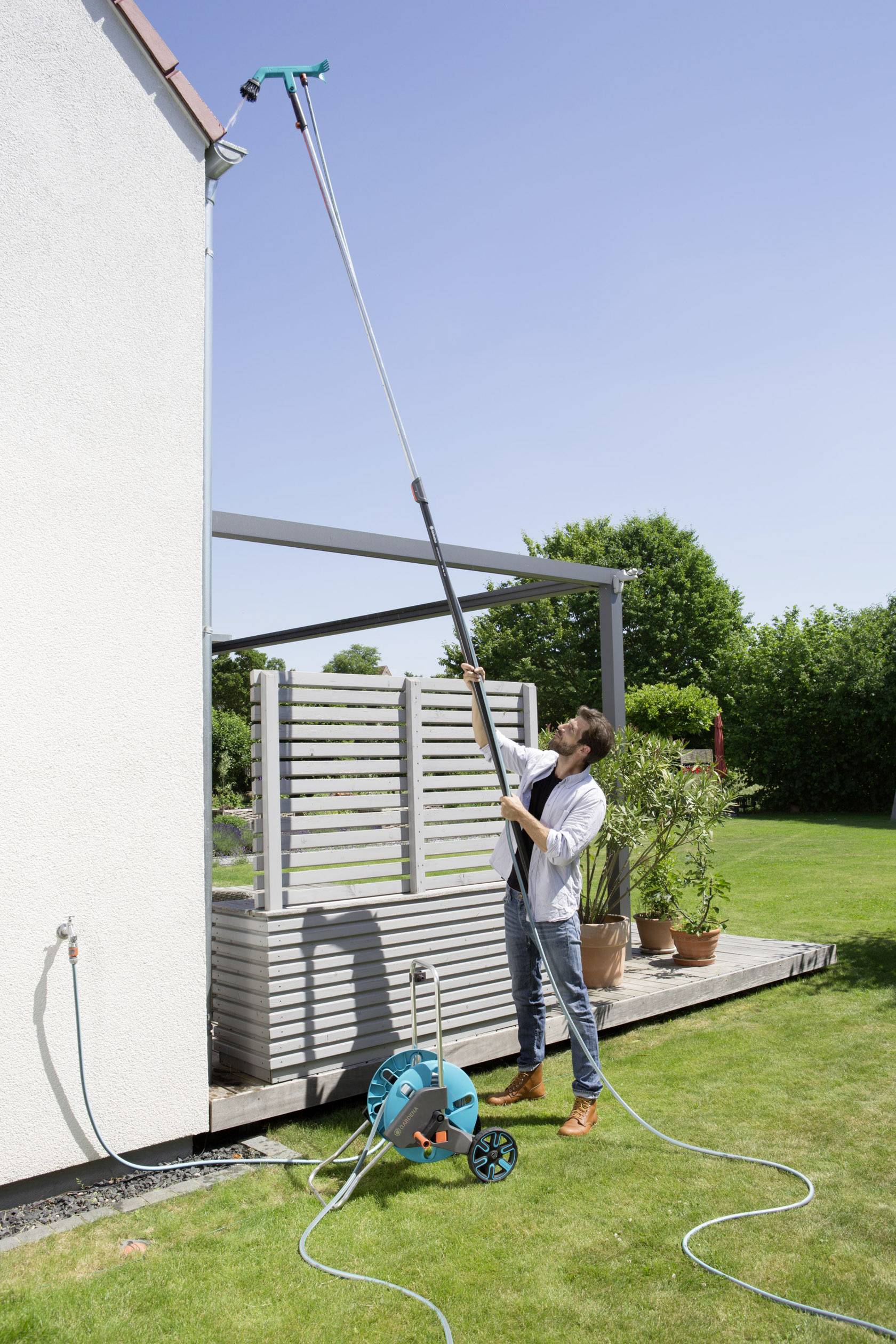 A person is using a long hose with a spray nozzle to clean the upper window of a house. The hose is connected to a water supply on the terrace.