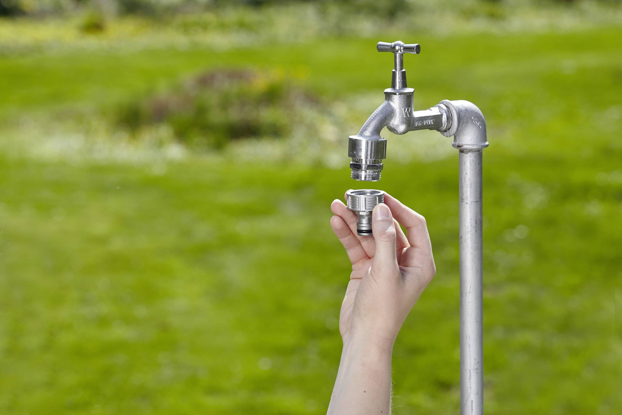A hand is holding a metal adapter in front of an outdoor tap, over green grass.