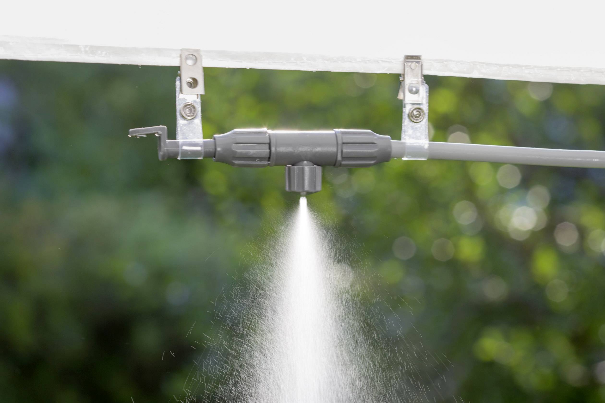 A grey sprinkler head of an irrigation system evenly distributes water in its surroundings. The background shows blurred greenery.