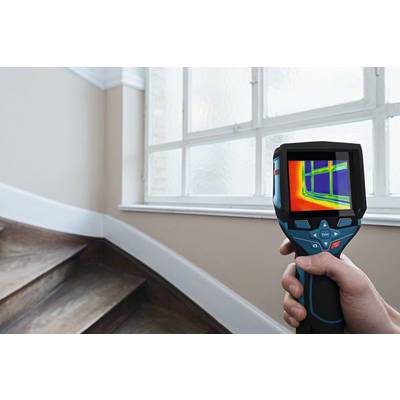 A person is holding a thermal imaging camera in front of a window. The screen displays temperature variations in the window area.