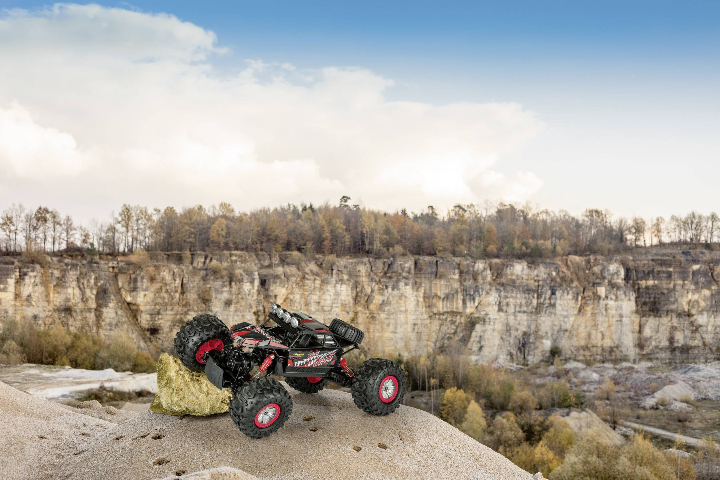 A remote-controlled car is positioned on a sandy slope in front of a rock face and wooded background under a blue sky.
