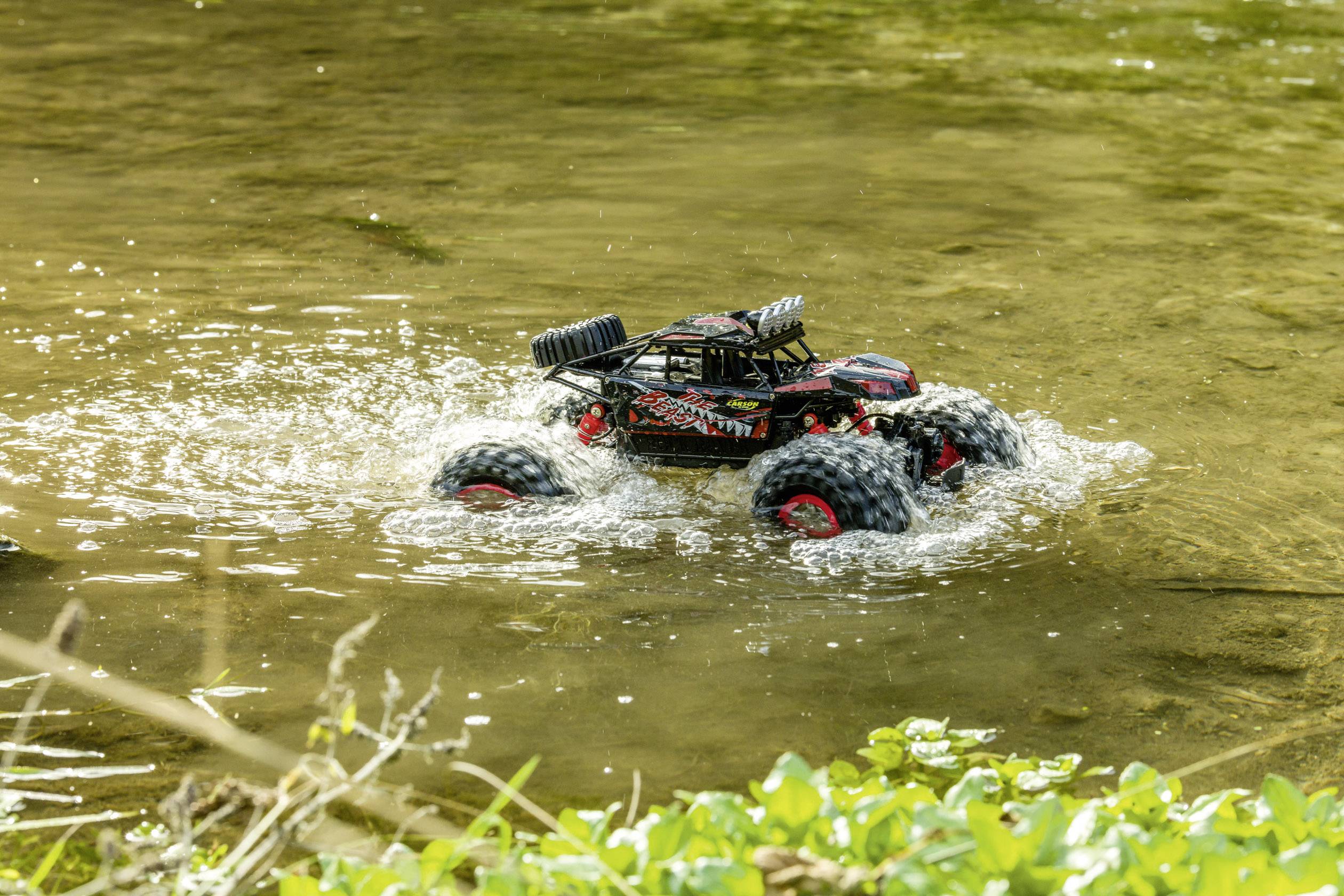 A black remote-controlled car with large tyres drives through shallow water in a natural environment.