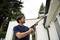 A person is cleaning the guttering of a house using a pressure washer. Trees and part of the house are visible in the background.