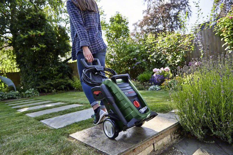A person is using a pressure washer on garden paving slabs. In the background, flowers are blooming and trees are towering upwards.
