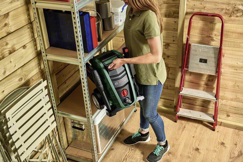 A woman in a green shirt places a green vacuum cleaner on shelves in a wooden shed. A ladder and folding chairs are in the background.