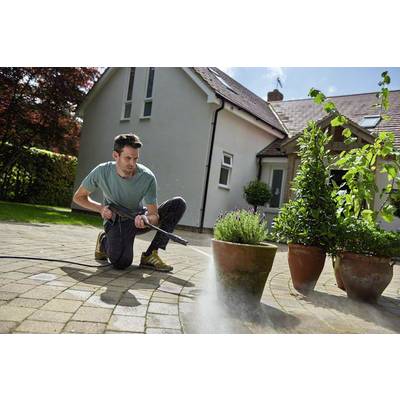 A man is cleaning a paved terrace with a pressure washer in front of a house, beside several potted plants.