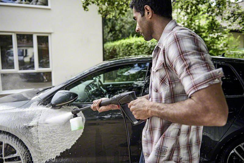 A man is washing a car with a pressure washer. Foam covers the front of the car. Green trees are in the background.