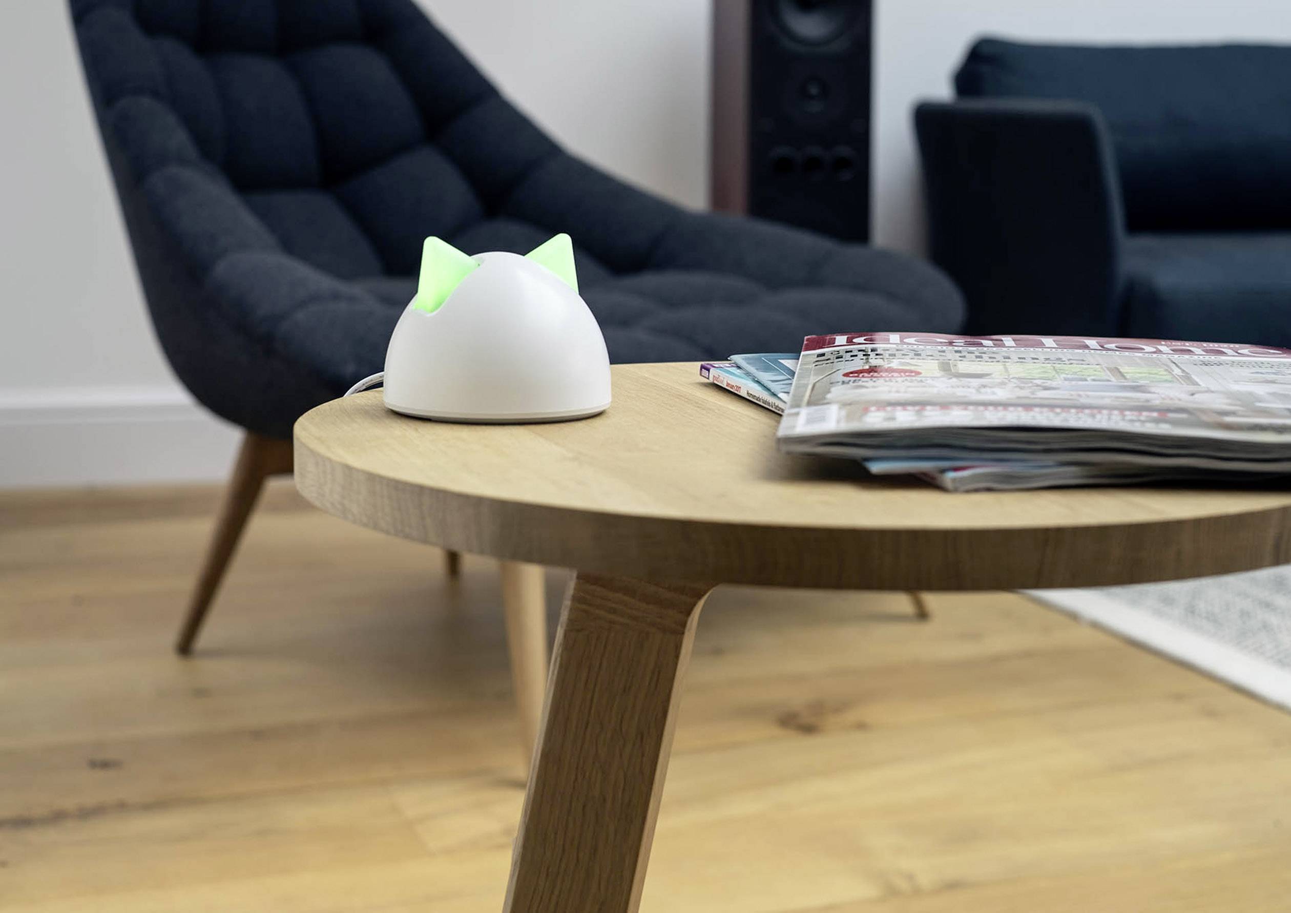 A contemporary living space with a round wooden table in the foreground. Magazines are placed on it and a device with green-glowing ears sits nearby. Two armchairs are positioned in the background.