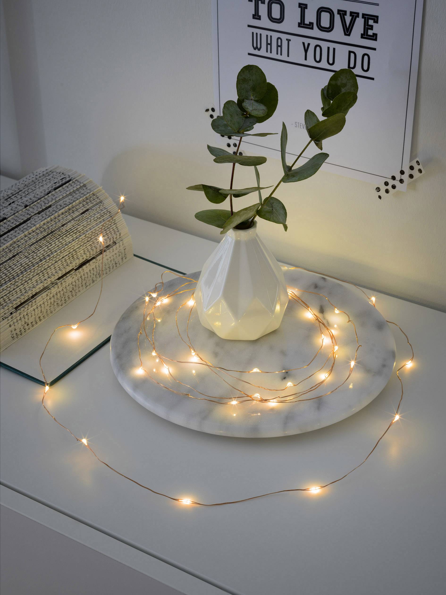 A small branch in a white vase on a table, surrounded by a string of fairy lights on a marble coaster, with a rolled-up carpet beside it.