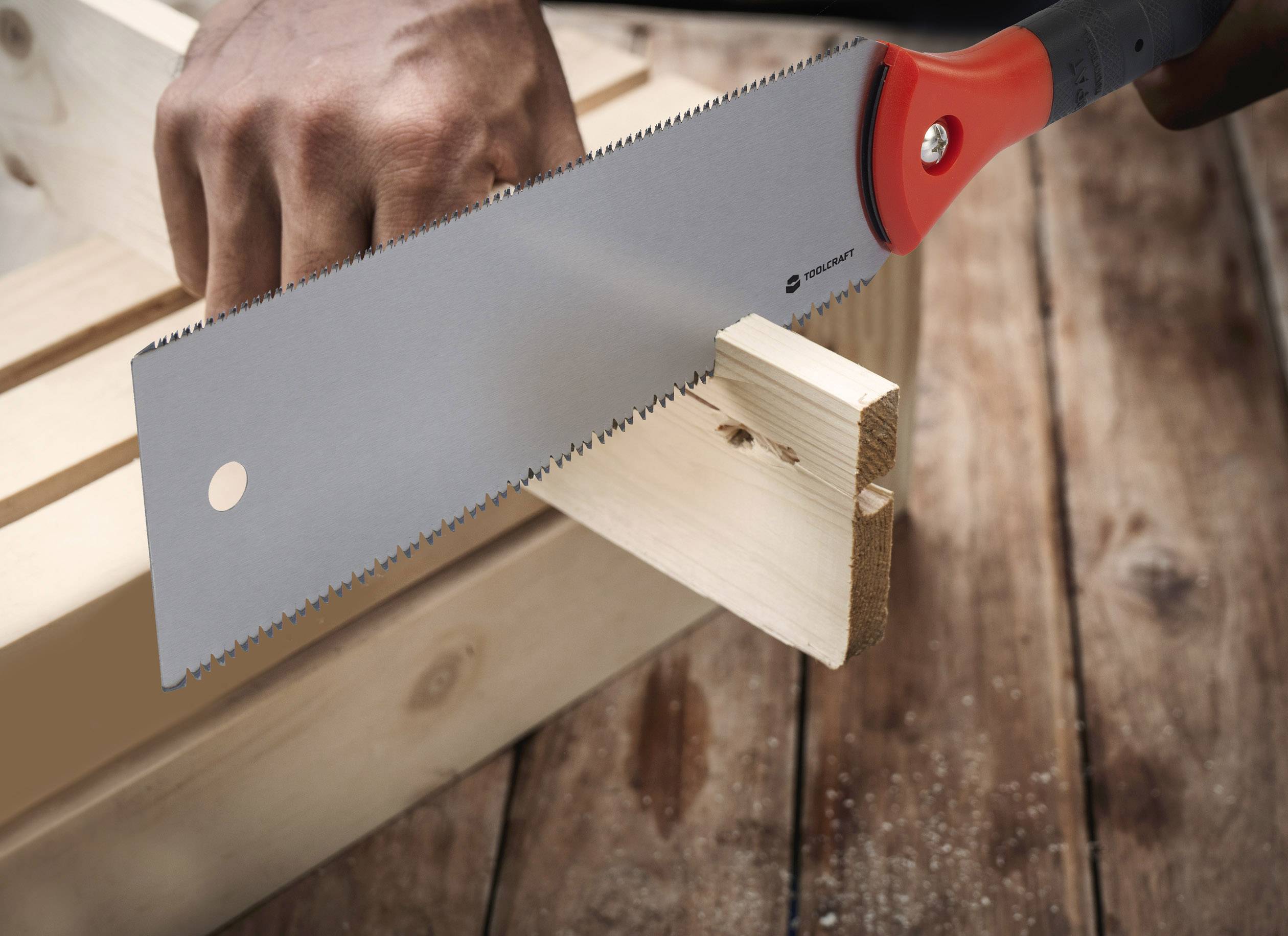 A person is sawing a wooden board with a handsaw. The saw is silver with an orange handle. The wood is lying on a table.