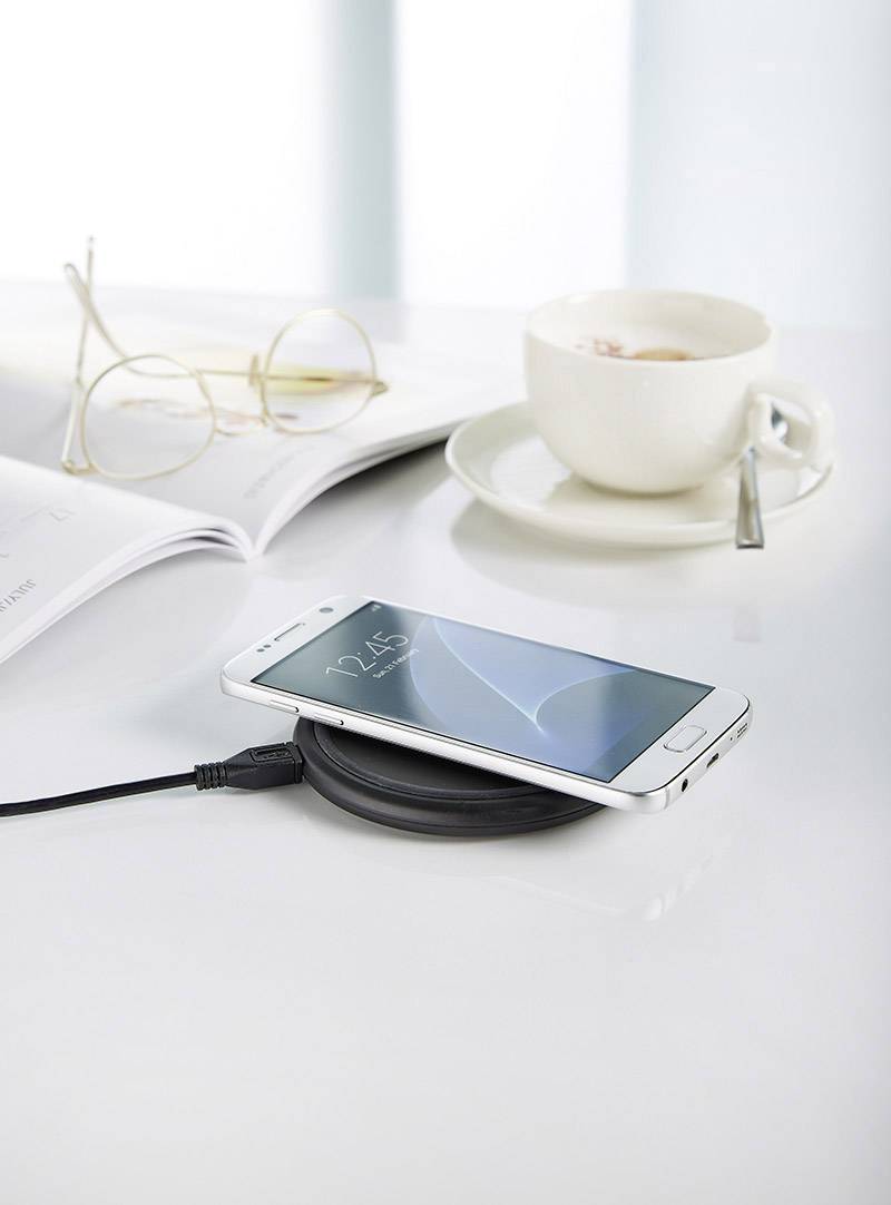 A smartphone rests on a wireless charger on a table. Beside it, an open pair of glasses, a book and a half-full cup of coffee.