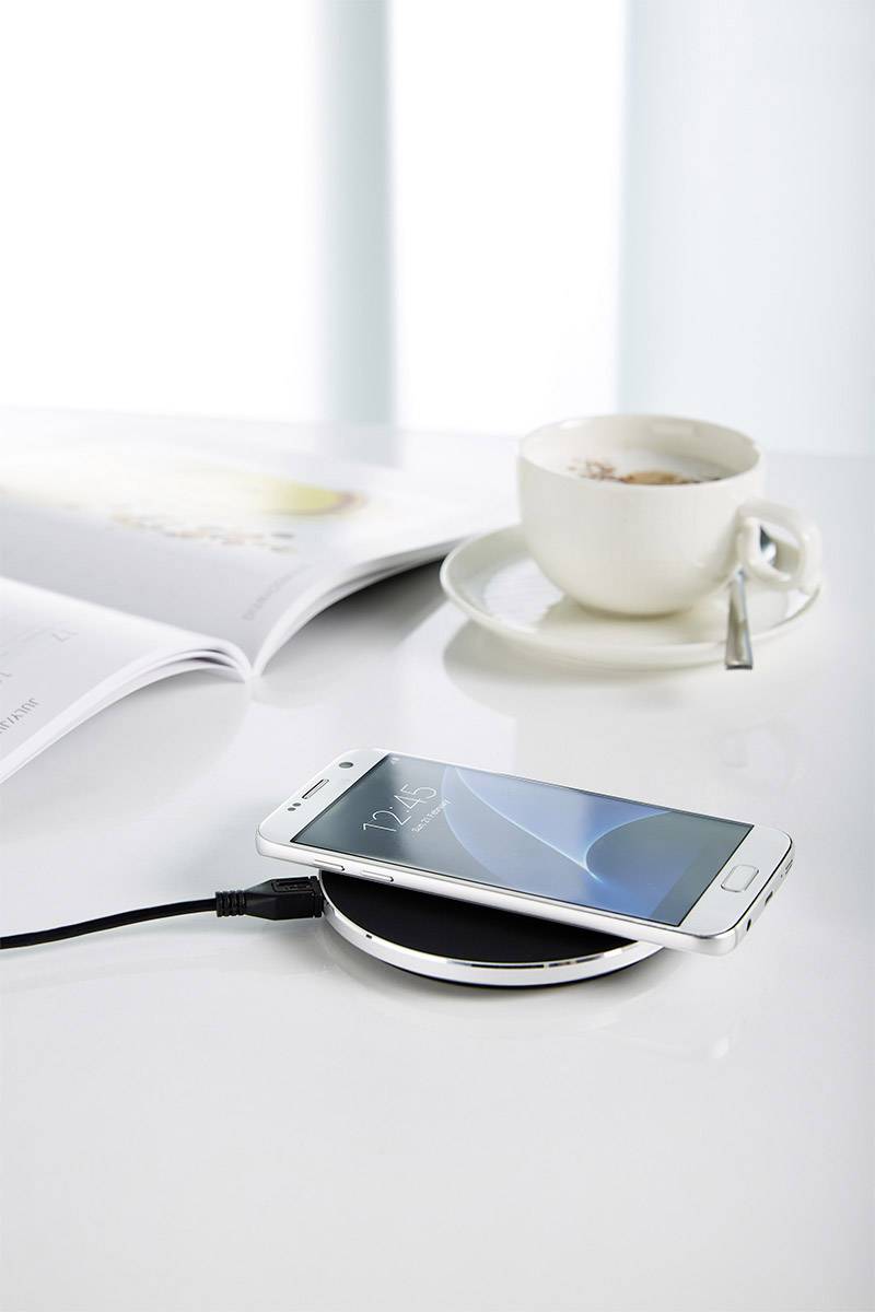 A smartphone is charging wirelessly on a table. Next to the phone are a coffee cup and an open magazine.