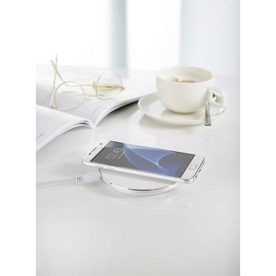 A mobile phone is charging wirelessly on a charging station. Beside it are a pair of glasses, an open magazine, and a cup of coffee.
