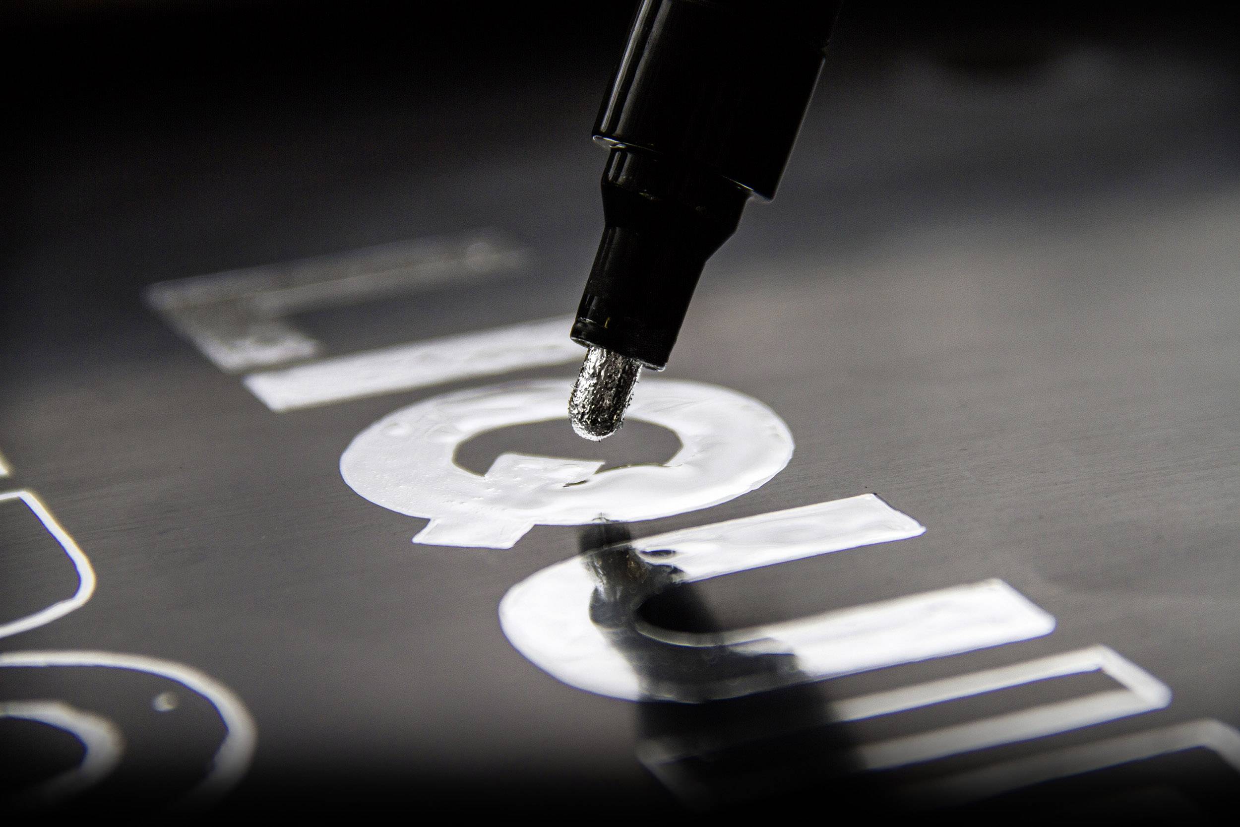 A thick black marker carefully fills in letters on a black surface, with the letter 'C' being highlighted.