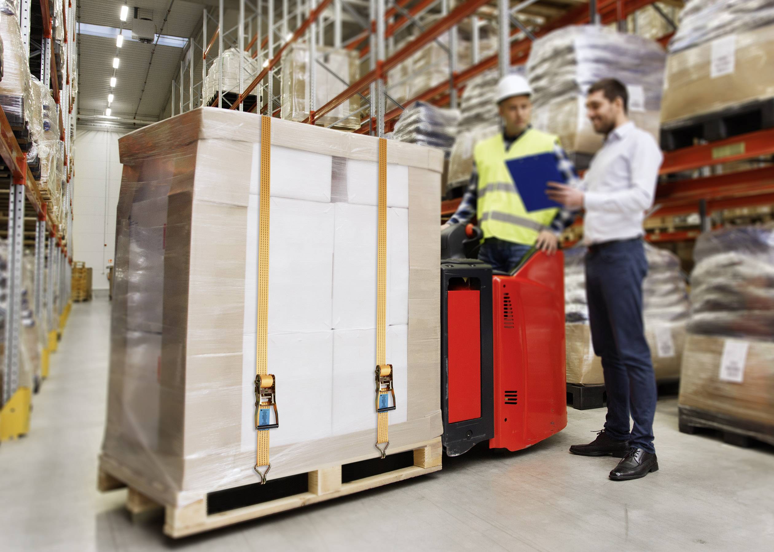 A warehouse worker in a high-visibility vest is driving a forklift with a pallet, while a second person shows him something on a clipboard.