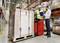 A warehouse worker in a yellow high-visibility vest operates a forklift with a pallet. A man is pointing at a clipboard.