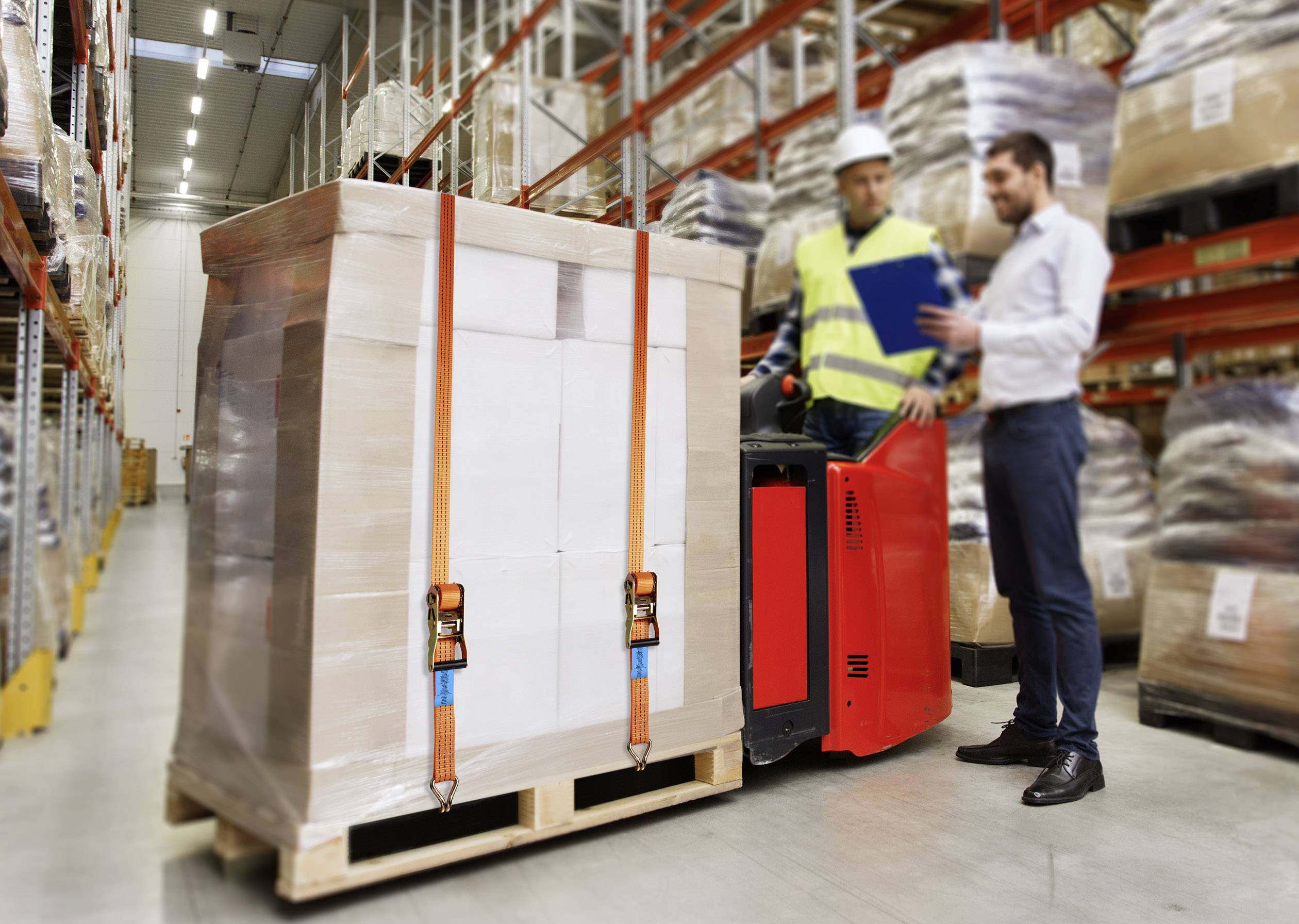 A warehouse operative is operating a forklift with a large load on a pallet. Another man is giving instructions in the warehouse.