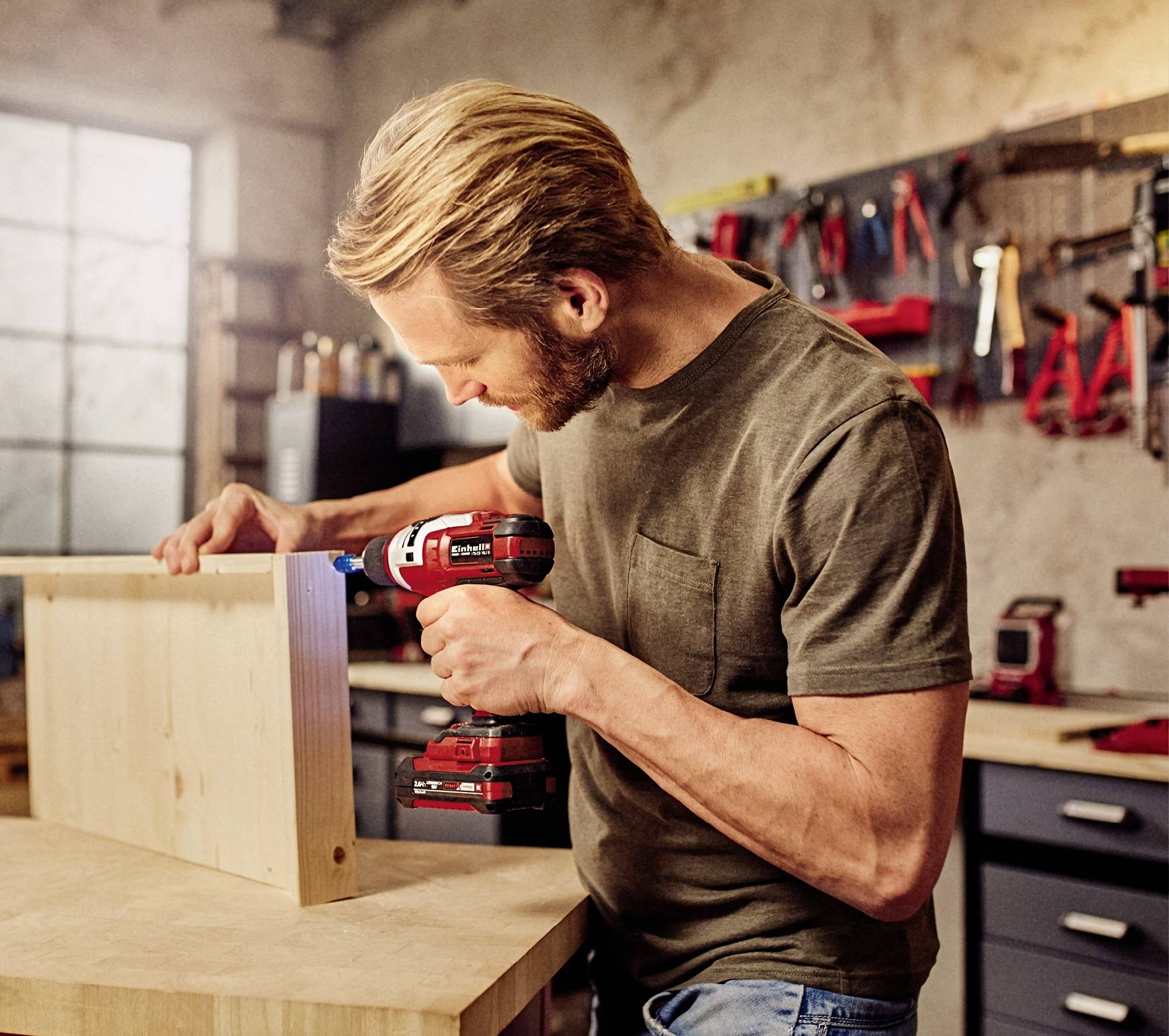 A man is installing a piece of wood using a cordless drill in a workshop, surrounded by tools and wooden furniture.