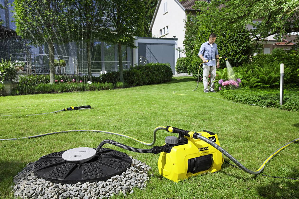 A man is watering a green lawn with a yellow garden implement. In the background, a lawn sprinkler is spraying water over the lawn.