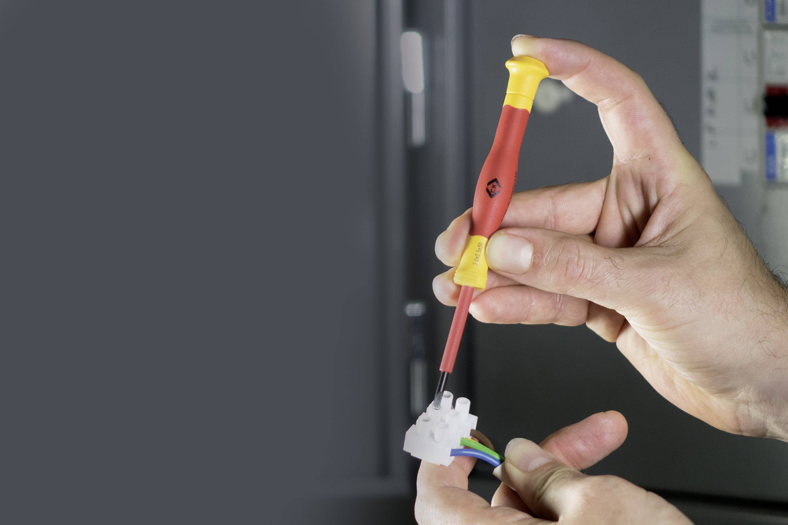 Two hands hold a screwdriver and a white plug. A control cabinet is visible in the background.