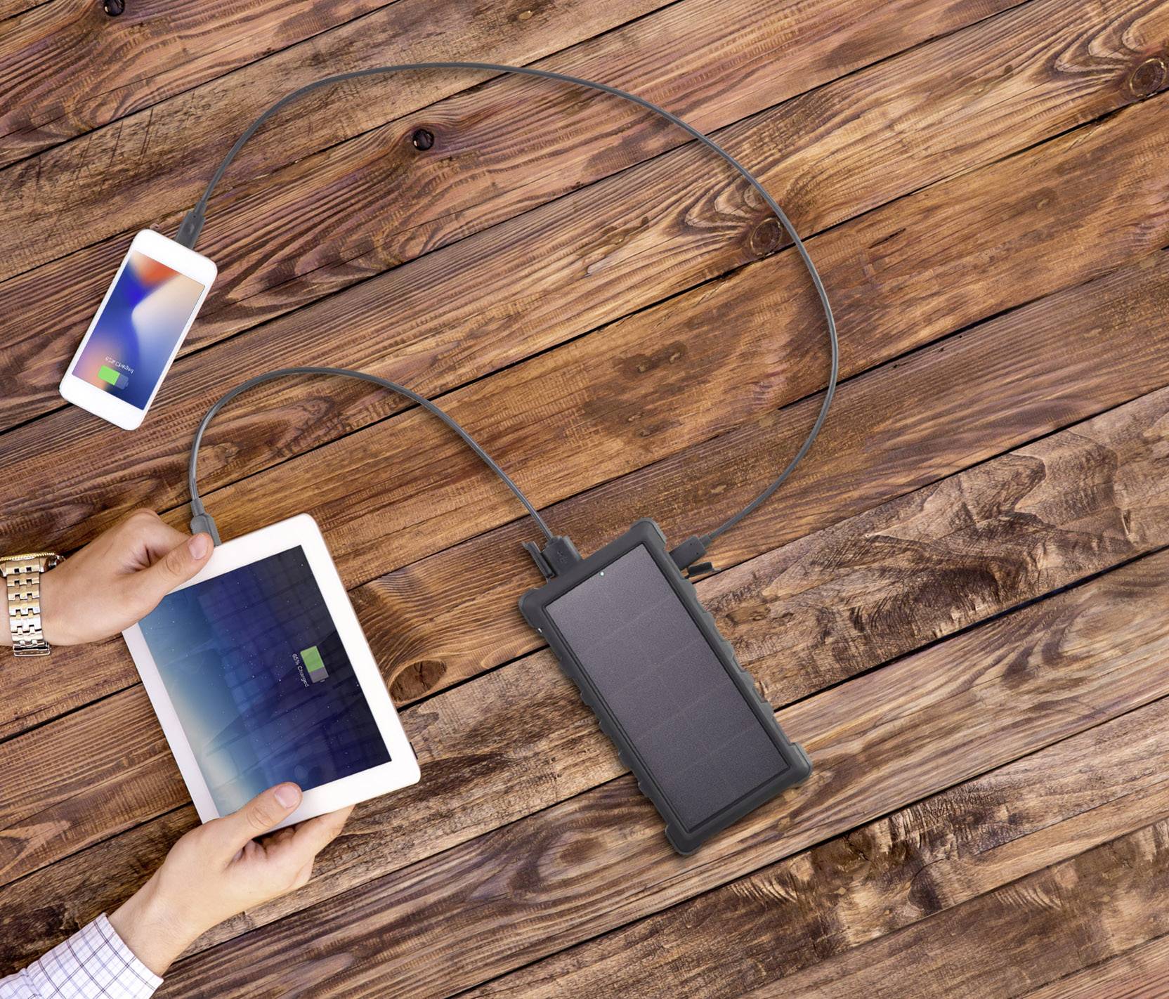 A solar charger is charging a smartphone and a tablet on a wooden table. The solar panel is connected to the devices via cables.