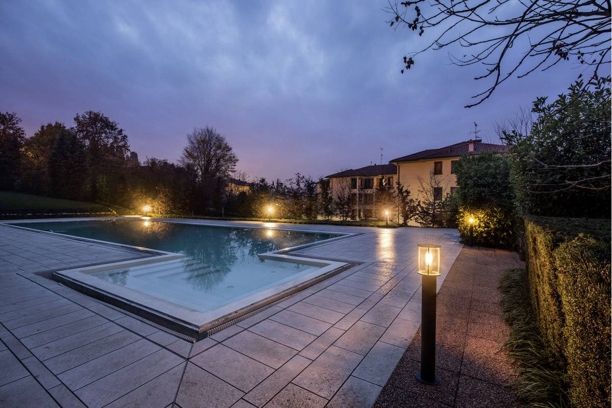 A illuminated outdoor swimming pool at dusk, surrounded by trees and with a house in the background, creates a tranquil atmosphere.