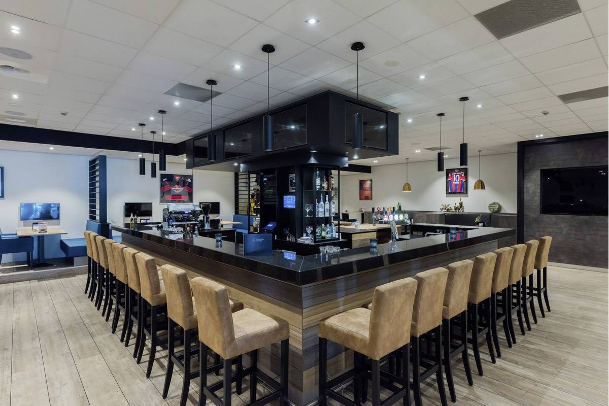 A modern hotel bar with leather bar stools around a large, rectangular counter. Screens and various decorations are situated in the background.