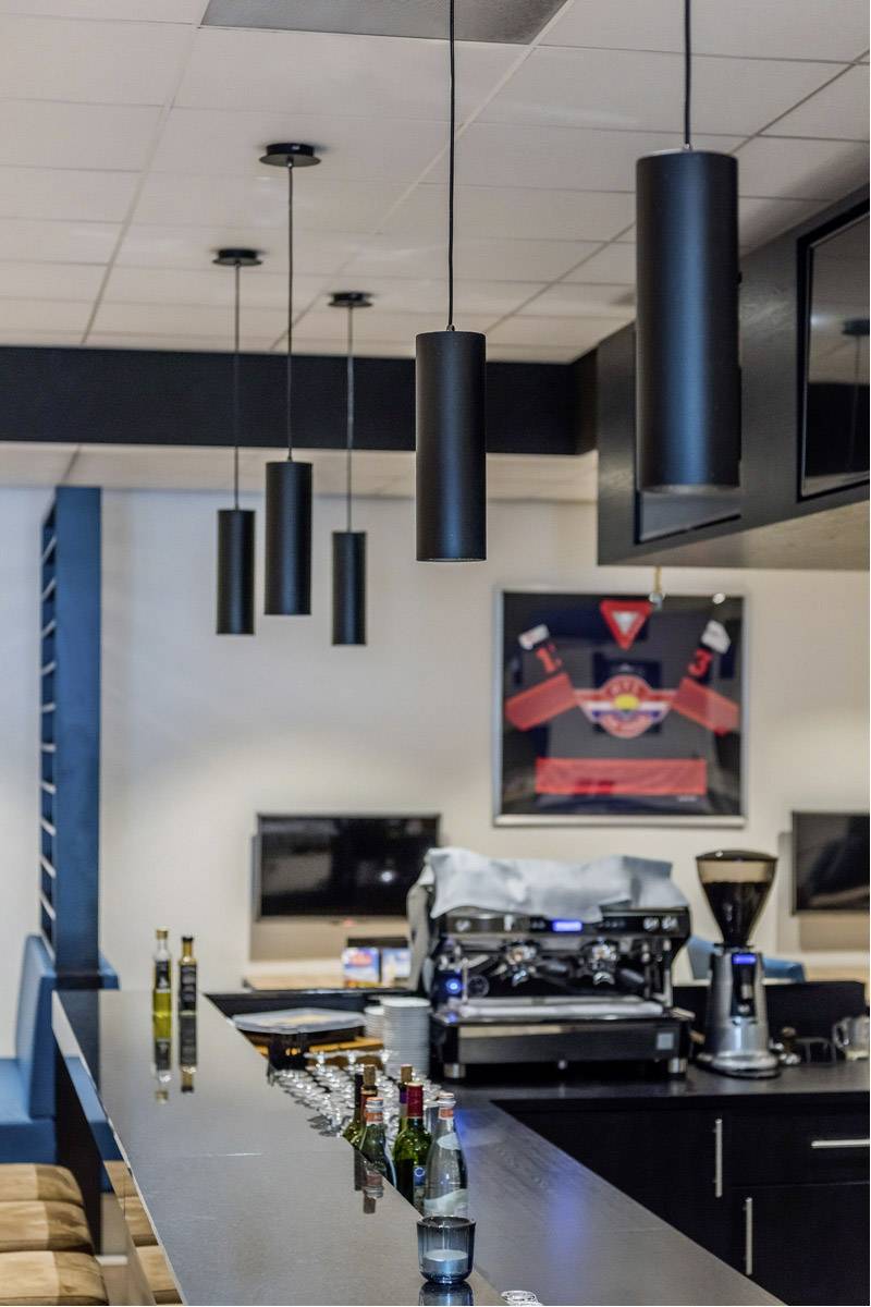 Modern bar area with a counter, hanging lamps, coffee machine, and drinks. A framed sports jersey hangs on the wall.