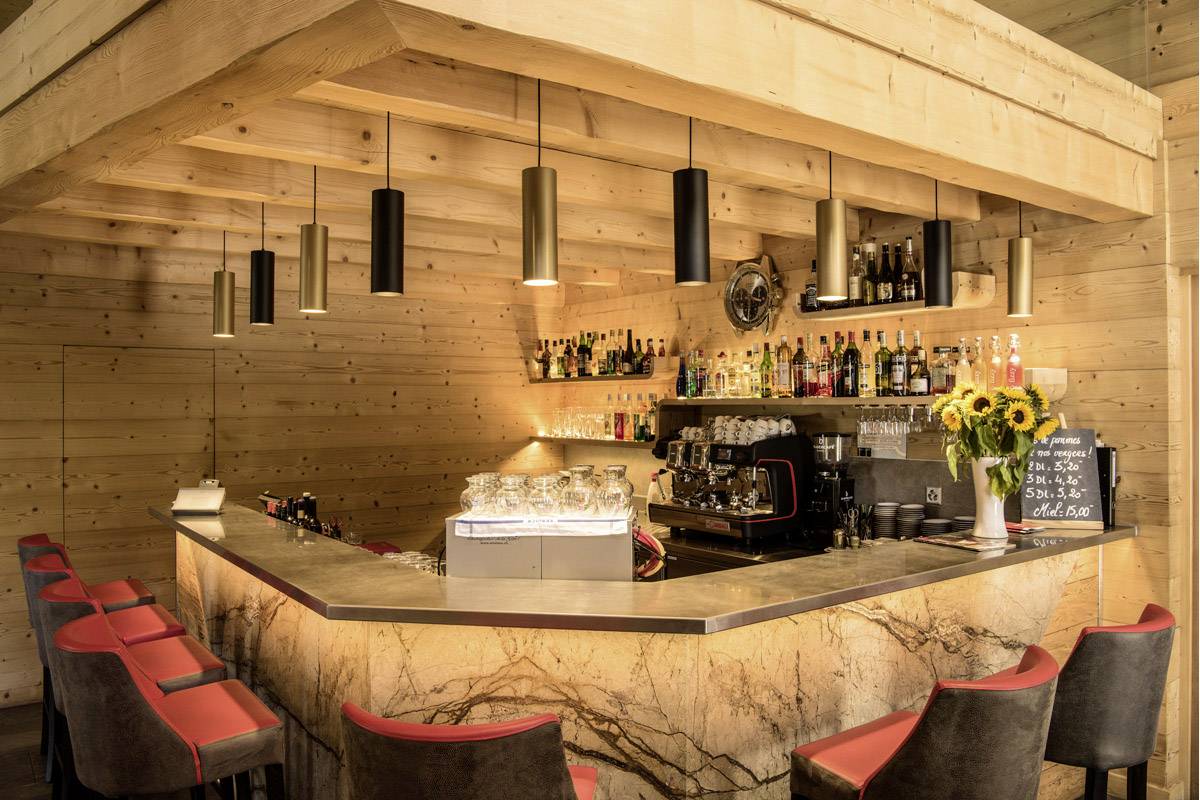A cosy bar with wooden walls, well-stocked with various drinks. Red bar stools and subtle lighting create a warm atmosphere.