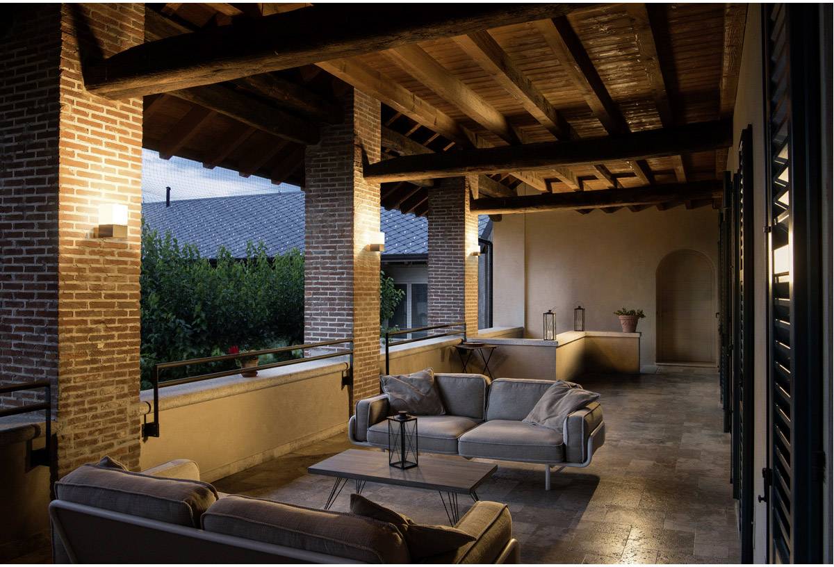 Covered terrace with cosy sofas and a table at dusk; brick walls and wooden beam ceiling lend a rustic charm.