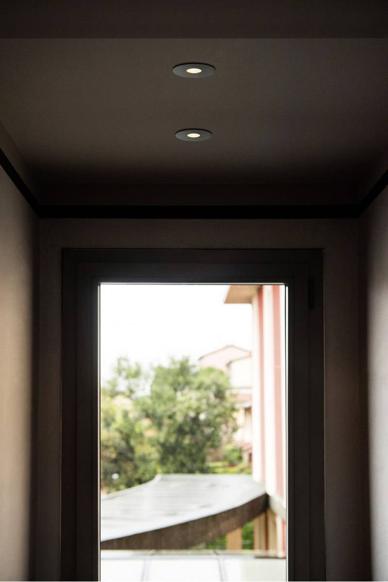 Hallway with a door, view outside to trees and a building. Ceiling lights are on.