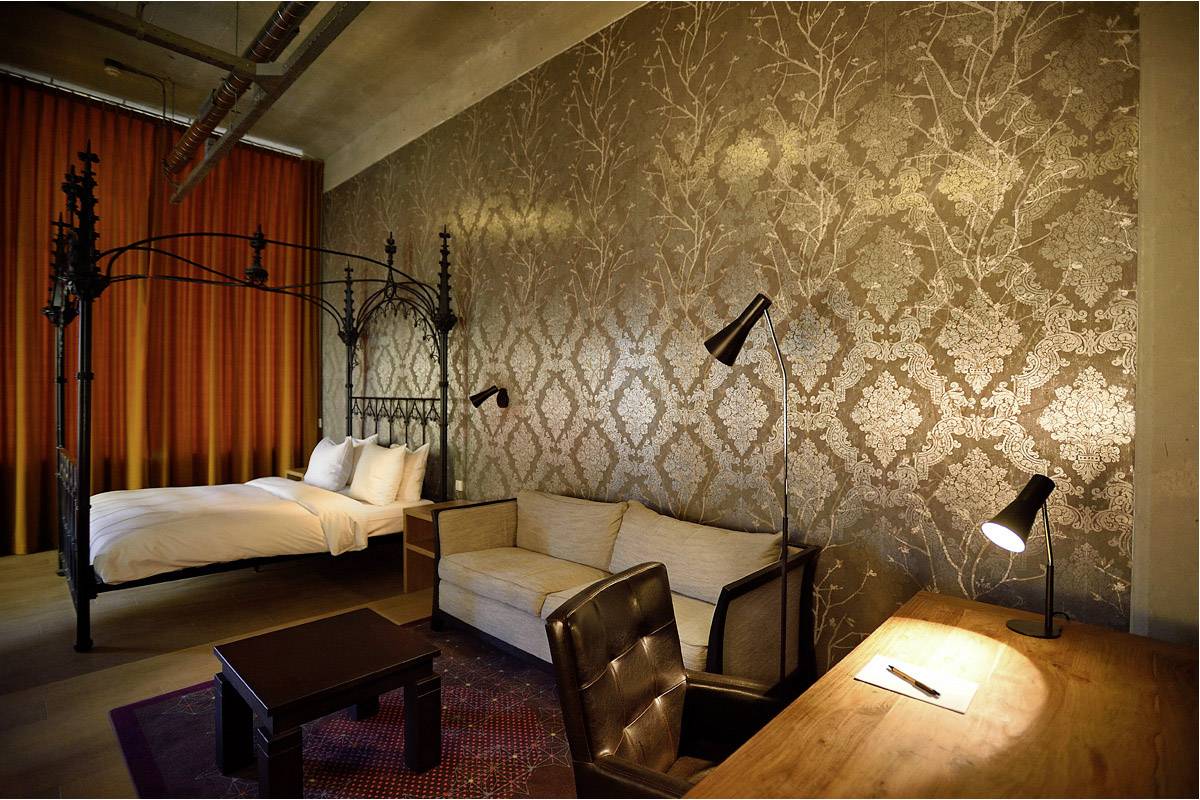 Hotel room with a four-poster bed, bronze patterned wallpaper, sofa, desk and standing lamps. Warm and elegant ambience.