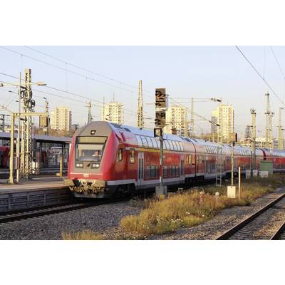 A red regional train travels along tracks in an urban railway station. High-rise buildings and electricity pylons are visible in the background.