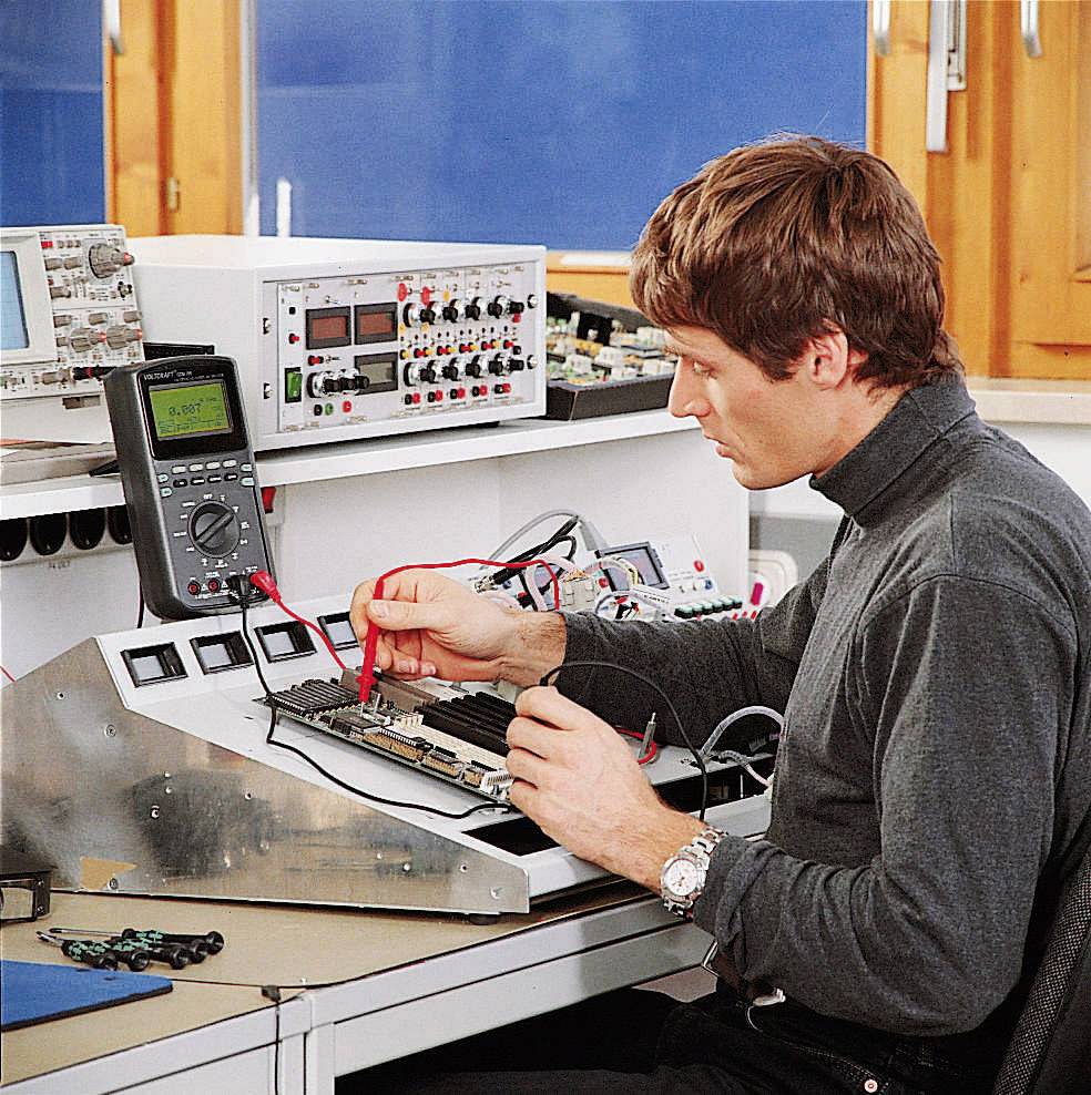 A man is working on electronic measuring instruments in a laboratory. He is using a multimeter to test circuits at a workbench.