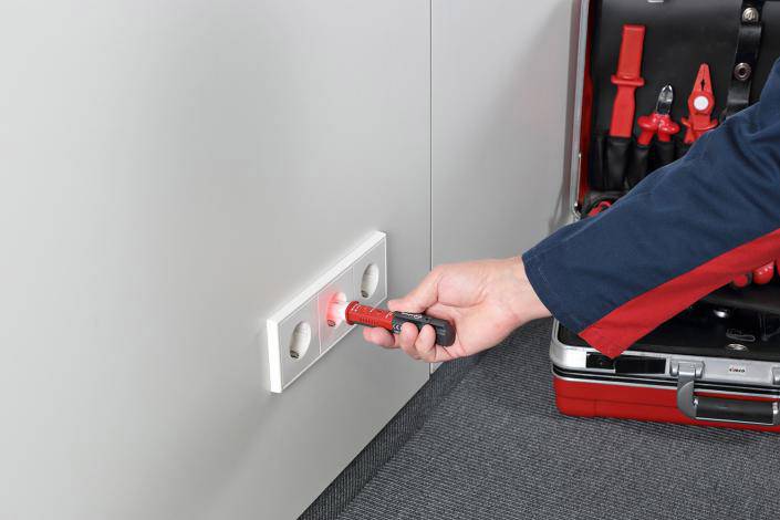 A person is testing a socket with a voltage tester. Next to the socket is an open toolbox with insulated tools.