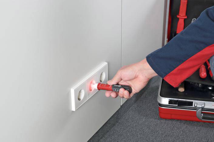 A person is checking a socket with a voltage tester. In the background, an open toolbox can be seen.