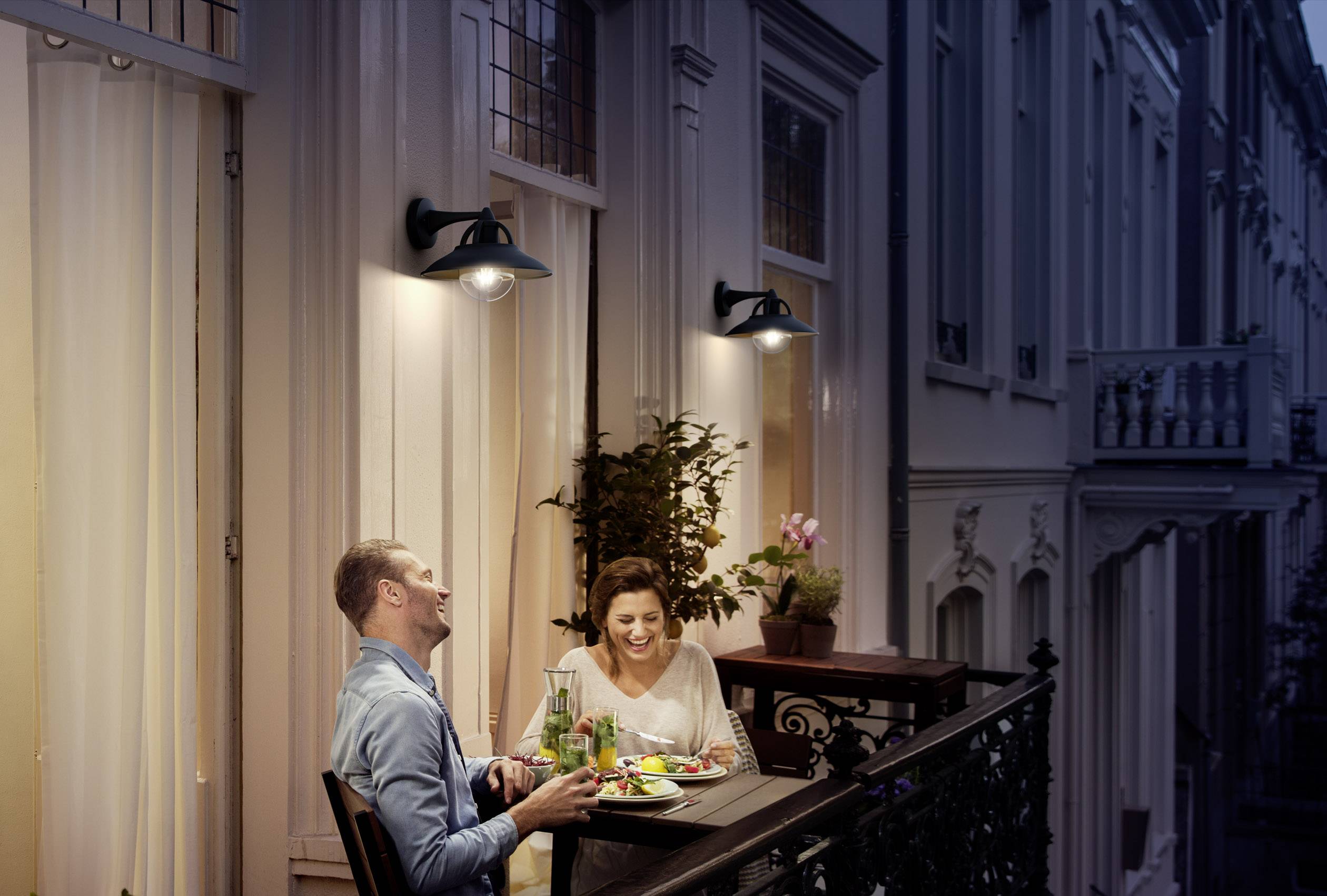 A couple sits on a balcony, eating dinner and laughing. The atmosphere is cosy, with warm lighting and a plant in the background.