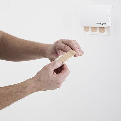A hand is sticking a plaster on a finger, while a dispenser with additional plasters hangs on the wall.