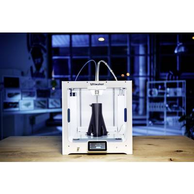 A 3D printer is printing a black object in a modern workshop environment. The printer has a white casing and a display.