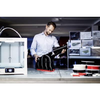 A man is examining a large 3D-printed object in a modern laboratory. Beside him stands a 3D printer and various filaments.